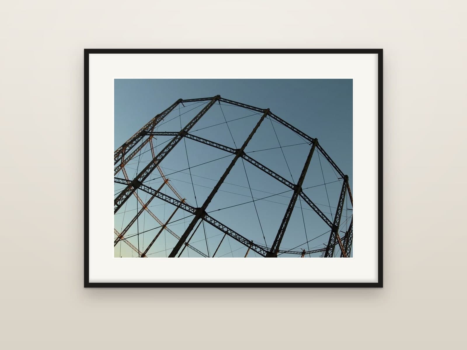 The iron lattice of a gasholder frame against a clear blue evening sky, shown as a framed print
