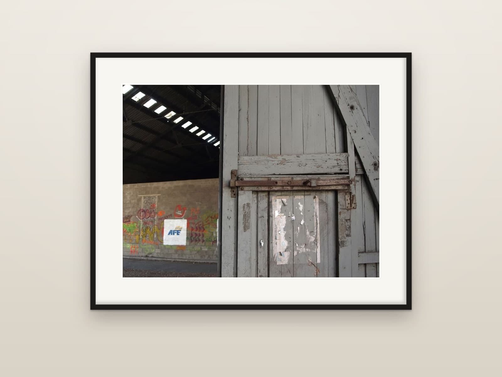 A worn wooden door with a rusted latch inside a railway shed with graffiti, shown as a framed print
