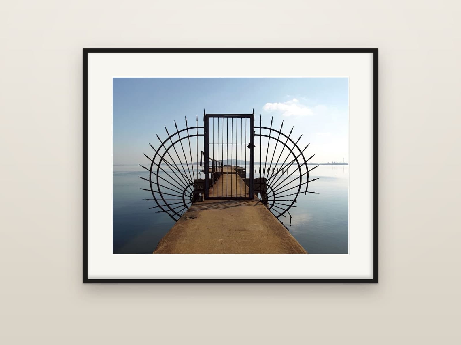 A spiked iron gate standing at the end of a pier over calm water, shown as a framed print